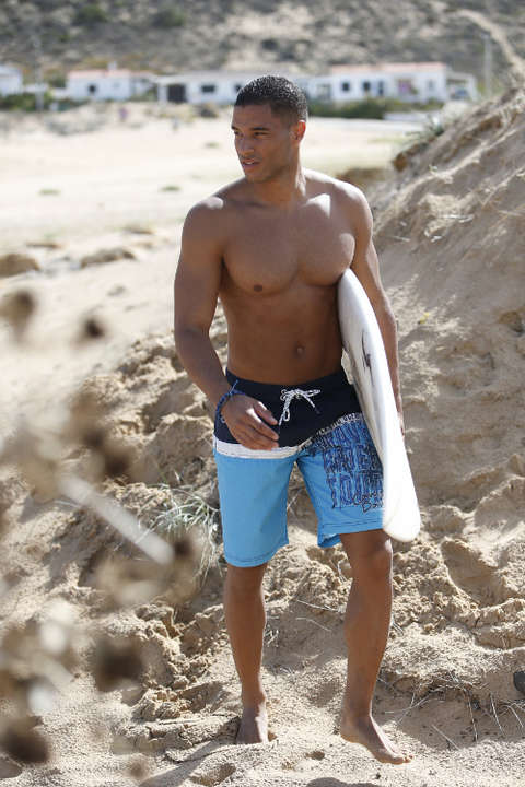 Ein Mann mit Surf-Badeshorts am Strand. Unter dem Arm trägt er ein Surfbrett.