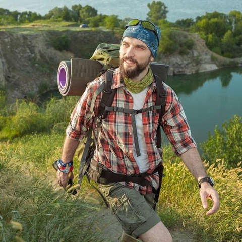 Ein Mann mit Bart wandert an einem See entlang. Er trägt ein kariertes Wanderhemd, Rucksack und Kopftuch.