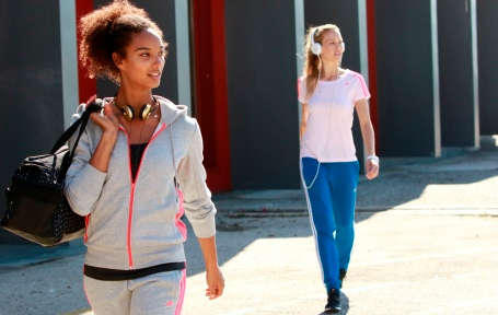 Im Vordergrund geht eine Frau in grauem Jogginganzug und einer Trainingstasche über der Schulter. Im Hintergrund geht eine weitere Frau mit blauer Jogginghose und rosa farbigen Oberteil.