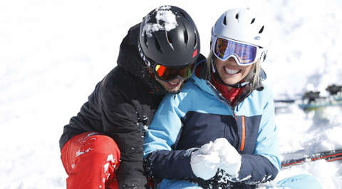 Ein Paar im Schnee bekleidet mit dicken Skijacken, Skihelmen und Skibrillen.