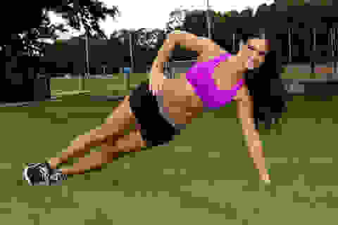 Eine Frau in kurzer schwarzer Hose und violettem Trainings-Top macht die Übung seitliche Planke auf einer Rasenfläche.