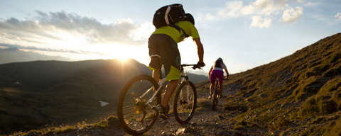 Zwei Mountainbiker fahren durch ein Gebirge. Am Horizont ist ein Sonnenuntergang zu sehen.