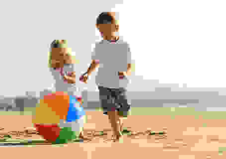 2 Kinder laufen barfuß am Strand und spielen mit einem Wasserball.