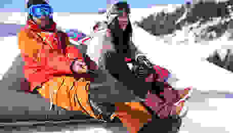 Ein Mann und eine Frau sitzen im Schnee. Der Mann zieht sich gerade seine Snowboardboots an.