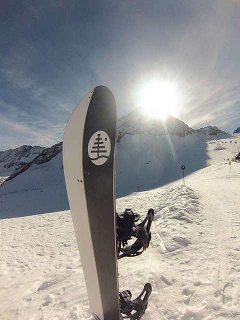 Ein Splitboard steckt hochkant im Schnee. Im Hintergrund ist ein Berg zu sehen, hinter dessen Gipfel, gerade die Sonne vorbei scheint.