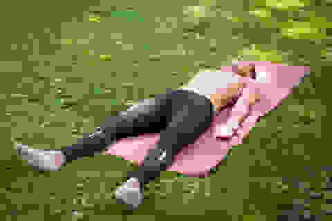 Ein Mann liegt mit freiem Oberkörper und schwarzer Hose auf einer Yogamatte im Park.