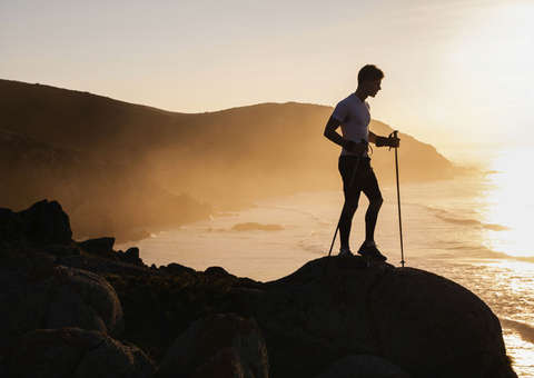 Ein Trailrunner steht mit Trailrunning Stöcken in der Hand während eines Sonnenuntergangs auf einer Anhöhe.