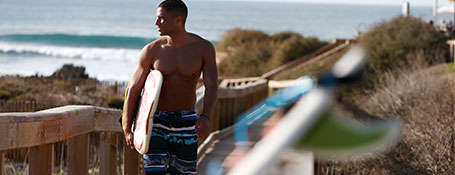 Ein Mann geht mit einem Surfbrett unter dem Arm einen Holzsteg am Strand entlang.