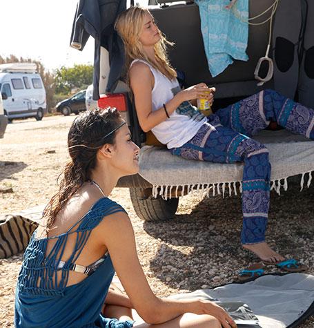 2 Frauen entspannen in der Sonne. Eine sitzt auf dem Boden, die andere im Kofferaum eines Autos.