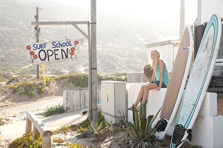 Eine Frau sitzt auf einer weißen Mauer vor einer Surfschule.