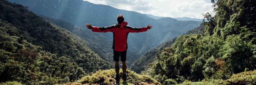 Ein Mann tr&auml;gt eine PFC freie Jacke von Jack Wolfskin. Er steht auf einem Berg und schaut mit zur Seite ausgebreiteten Armen in das gr&uuml;ne Tal.