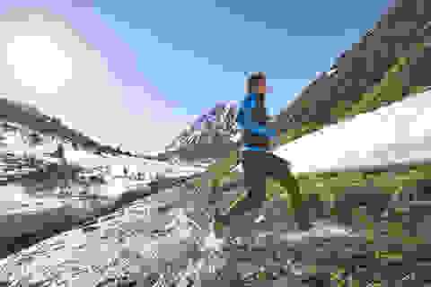 Ein Trailrunner läuft durch einen kleinen Bach. Im Hintergrund ist ein schönes Bergpanorama zu sehen. 