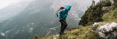Ein Wanderer geht bei Regen über einen Wanderweg während er eine türkise Wanderjacke trägt