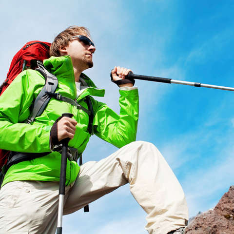 Ein Wanderer in grüner Wanderjacke mit rotem Rucksack, Sonnenbrille und Wanderstöcken erklimmt einen Gebirgsweg.
