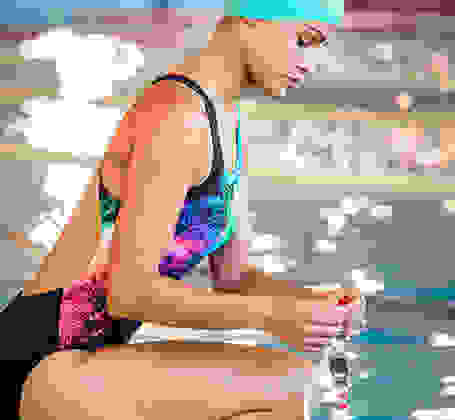 Eine Frau kniet im Badeanzug an einem Schwimmbecken und hält ihre Schwimmbrille in den Händen.