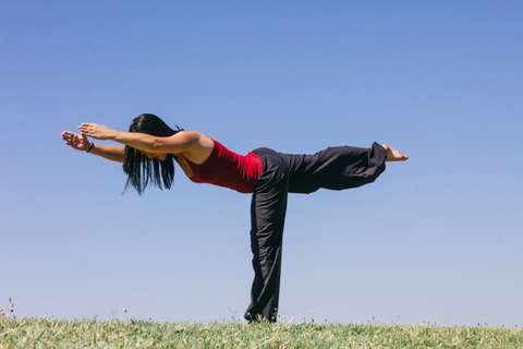 Eine Frau in schwarzer Hose und rotem Oberteil macht eine Yoga Übung unter freiem Himmel.